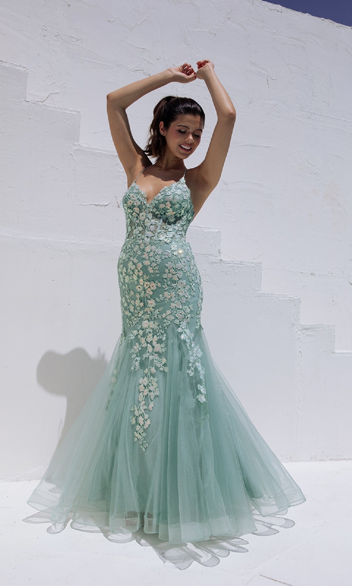 A woman in the Sequin-Floral Long Mermaid Prom Dress: Eureka 9696 stands outdoors against a white textured wall, raising her arms and smiling.